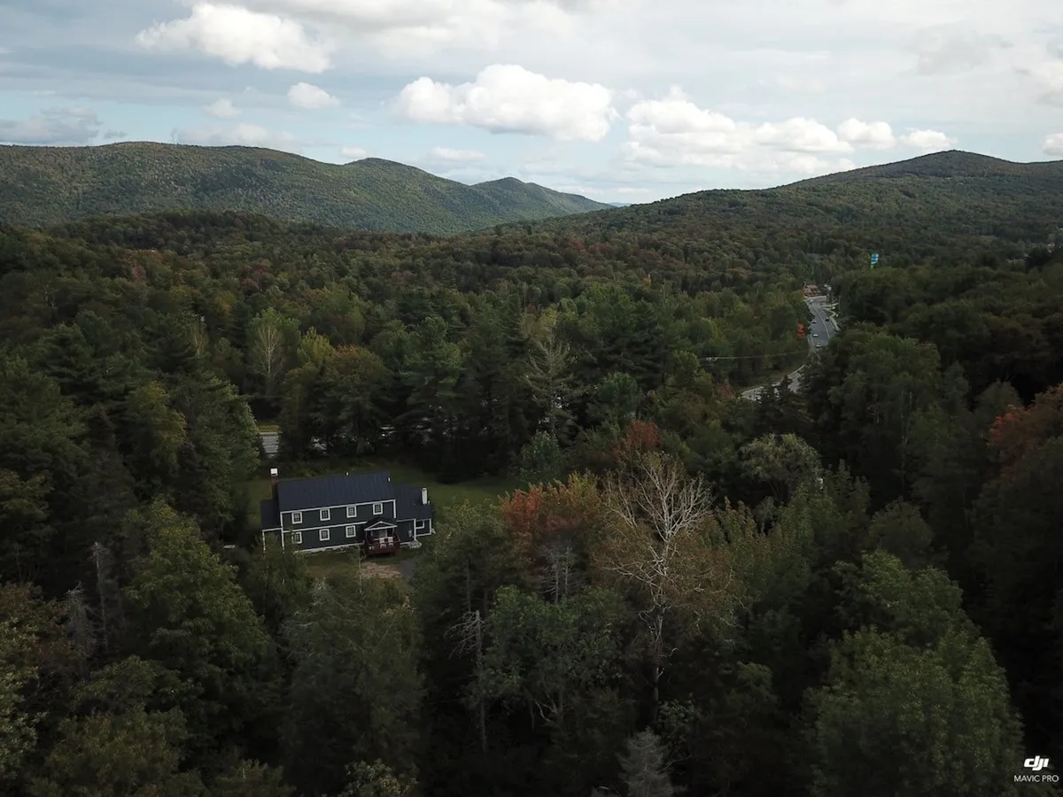 A wide view of the Bigelow Chalet nestled among tall trees, with distant mountains rising in the background. The porch is visible, and the setting sun casts a warm glow over the landscape, evoking a sense of tranquility and adventure. The natural environment hints at the proximity to hiking trails, ski slopes, and Vermont’s natural beauty.
