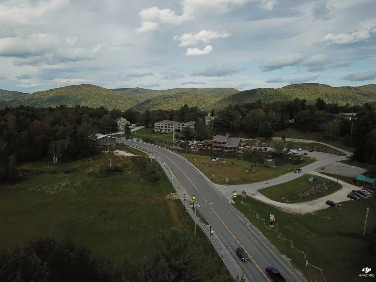 A panoramic view of rolling mountains, a winding road, and small local businesses nestled in the valley. The scene suggests easy access to both natural wonders and local culture, with hints of nearby restaurants and nightlife.