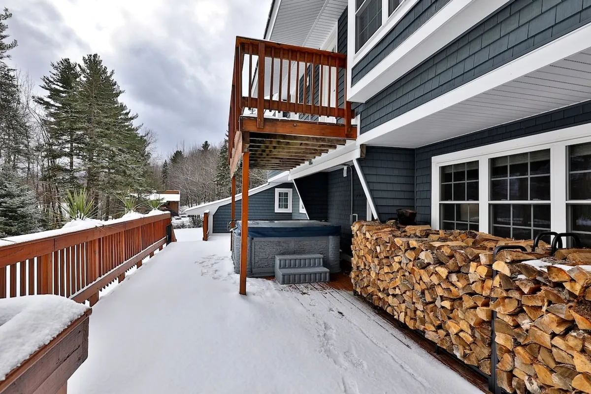 A snow-covered wooden deck featuring a bubbling hot tub and a tidy stack of firewood. The surrounding trees create a sense of privacy and immersion in nature, while the winter setting highlights the inviting warmth of the hot tub after a day on the slopes.
