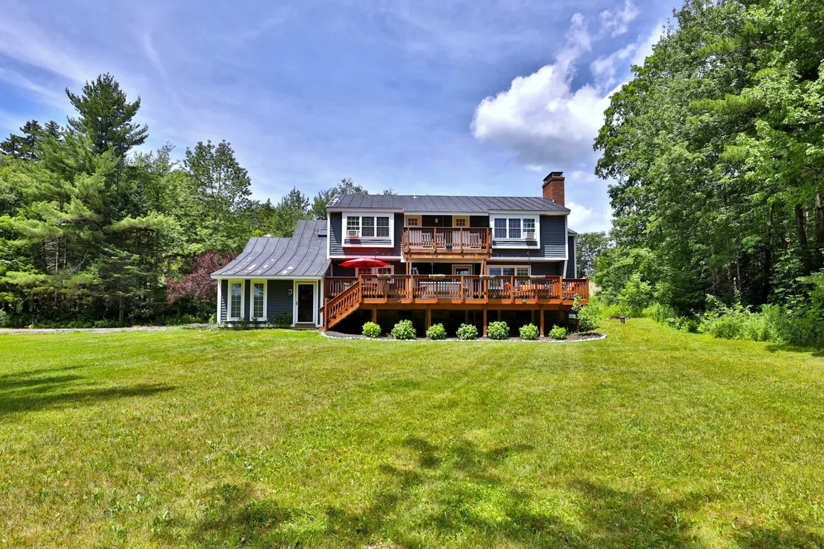 A broad view of a large wooden deck with a red umbrella, overlooking a green backyard lawn bordered by mature trees. The space is set up for gatherings, lawn games, or pet playtime, exuding a serene and welcoming atmosphere.