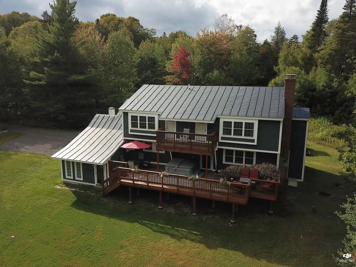 The image showcases the exterior of the Bigelow Chalet, featuring a modern roofline, a spacious wooden deck perfect for gatherings, and lush Vermont trees glowing with autumn colors all around. The setting evokes a tranquil mountain retreat, with the deck inviting guests to enjoy sunset views, morning coffee, or evening bonfires.