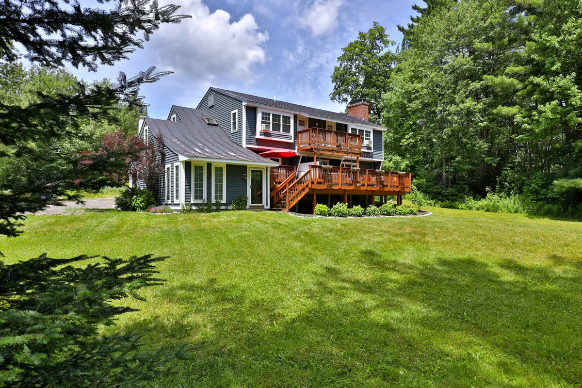 A wide view of the chalet's large wooden deck and lush green lawn surrounded by mature trees, with plenty of space for outdoor play and gatherings. The house exterior and deck seating are visible, creating an inviting atmosphere for family activities and relaxation.