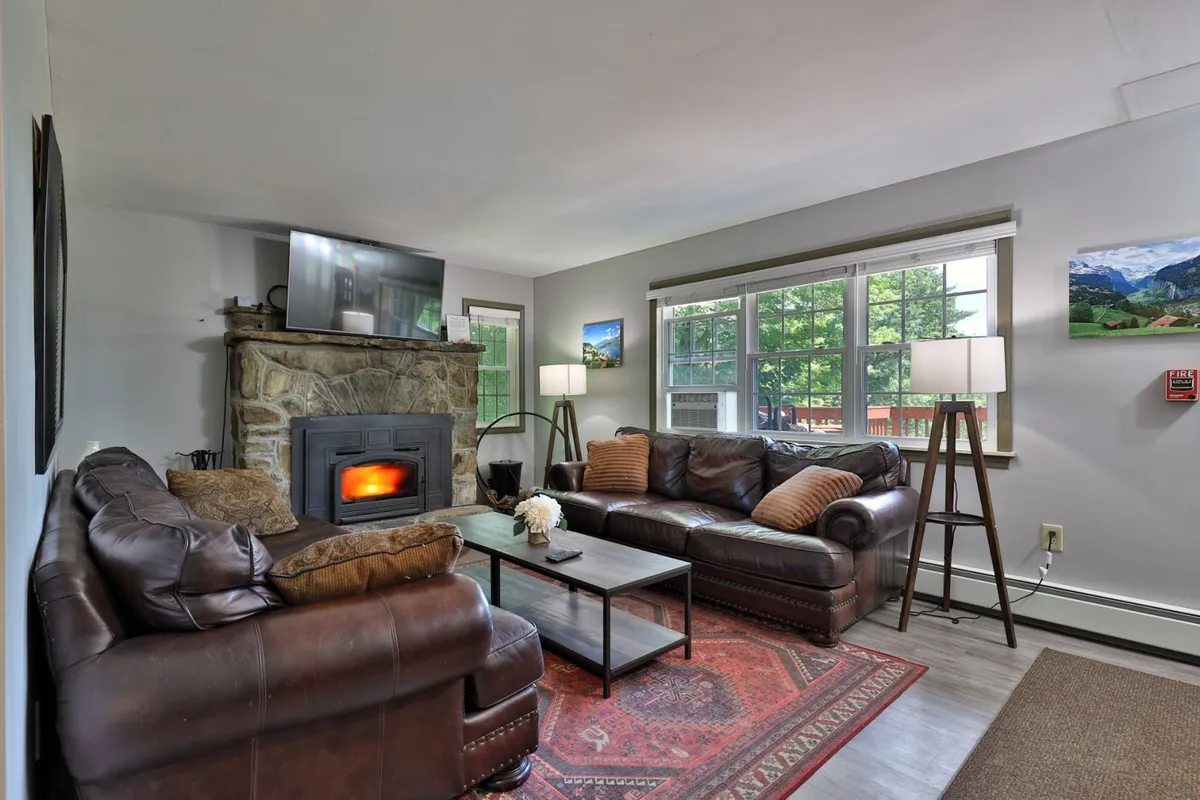 A cozy main living room with a prominent stone fireplace as the focal point. Plush sofas are arranged around a central coffee table, and large windows flood the space with natural light. The layout encourages group relaxation, storytelling, and watching TV together.
