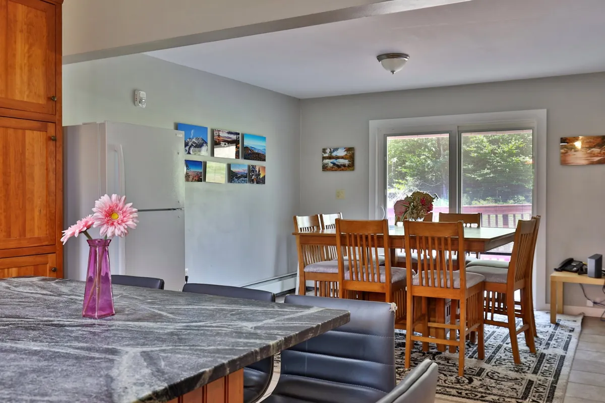 This inviting interior view captures a kitchen island that seamlessly transitions into a spacious dining area. The dining table, adorned with a flower vase and surrounded by ample seating, is set for a large group, suggesting a comfortable and social atmosphere for shared meals.