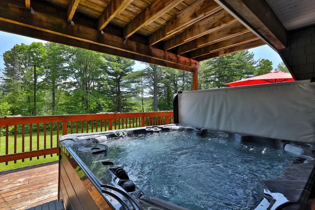 A tranquil exterior view of a wooden deck featuring a spacious six-person hot tub, surrounded by lush Vermont greenery. The scene shows people relaxing in the hot tub, socializing, and enjoying nature, with hints of the deck being suitable for gatherings.