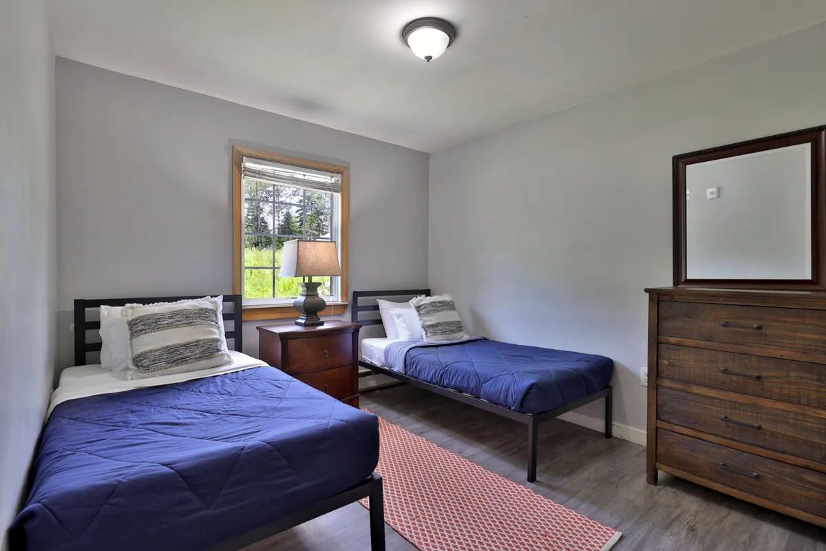 A bright bedroom with two single beds, a wooden dresser, and a window that lets in plenty of sunlight. The nightstand and lamp create a cozy, welcoming setting ideal for families, friends, or children sharing a room.